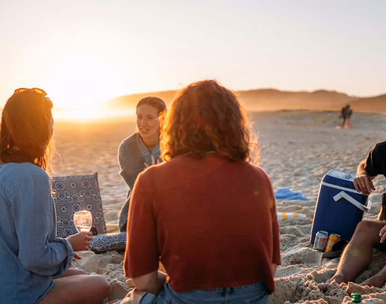 A group of friends on a sandy beach with the sun setting in the background talking